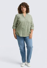 Camicia verde chiaro con bottoni, tessuto testurizzato e maniche arrotolate, abbinata a jeans blu a gamba dritta e sneaker bianche.