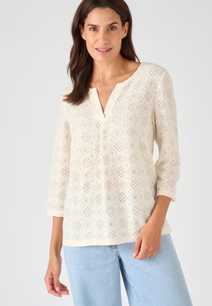 Blusa in pizzo bianco con aperture a forma di diamante, maniche a 3/4 e scollo a V. Abbinata a jeans denim celeste chiaro. Tessuto con design testurizzato.