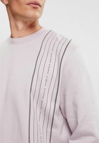 Sudadera rosa claro con cuello redondo, que presenta rayas verticales negras y texto a lo largo del lado izquierdo. Tejido suave, ajuste relajado.