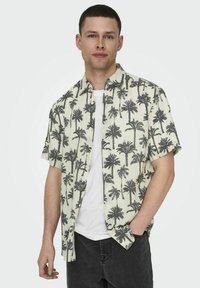 Chemise à manches courtes de couleur crème clair avec un imprimé de palmiers noirs, dotée d'un col et d'une fermeture à boutons. Portée par-dessus un t-shirt blanc.