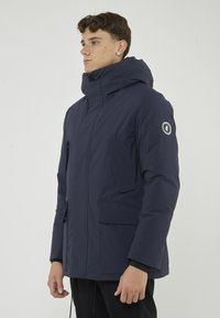 Giacca imbottita blu navy con colletto alto e cappuccio removibile, dotata di diversi taschini frontali e una patch con logo sulla manica. Texture liscia.