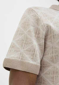 Beige pletený svetr se strukturálním geometrickým vzorem a žebrovanými manžetami, který zdůrazňuje jemný materiál a elegantní design.