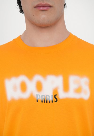 Oranžs kokvilnas T-krekls ar uzrakstu "KOOPLES" izplūdušā baltā fontā un "PARIS" treknajos melnajos burtiem, apaļa apkakle, maiga tekstūra.