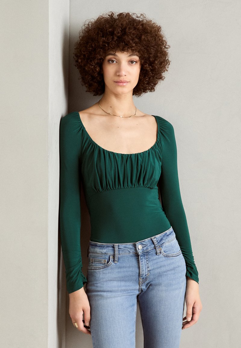Mørkegrønn bodysuit med lange ermer, scoop hals og rynket midje. Kombinert med lyseblå tettsittende jeans. Glatt stoff med subtil glans.