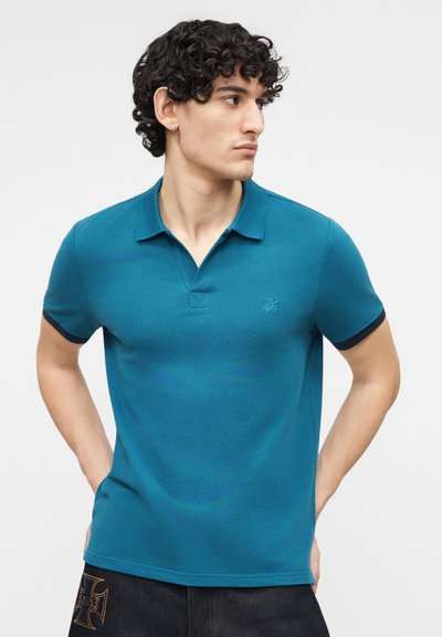 Νεαρός άνδρας με μπούκλες, φορώντας μια τουρκουάζ πόλο μπλούζα και σκούρο παντελόνι, κοιτάζοντας προς τα δεξιά με τα χέρια πίσω από την πλάτη.