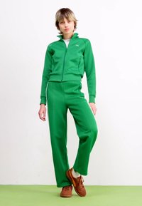 Grøn atletisk tracksuit lavet af et glat stof med en jakke med lynlås og bukser med elastisk talje. Brune sneakers fuldender outfittet.