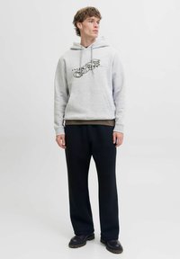 Młody mężczyzna w jasnoszarej hoodie z graficznym logo, czarnych spodniach i czarnych butach, stojący na tle gładkiego białego tła.