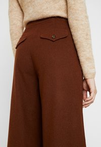Beige lange mouwtrui gestopt in hoge taille, wijde bruine broek met twee knoopzakken aan de achterkant, weergegeven van achteren.
