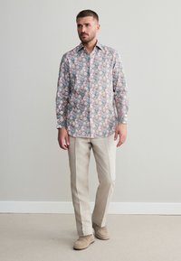 Bloemenblouse met lange mouwen met lichtblauwe, roze en crèmekleurige bloemen op een zachte grijze achtergrond; gecombineerd met beige broek en lichte schoenen.
