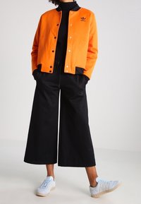 Orange bomberjacka i bomull med tryckknappar, svarta vida byxor och ljusblå sneakers med vita detaljer. Svart polotröja under.