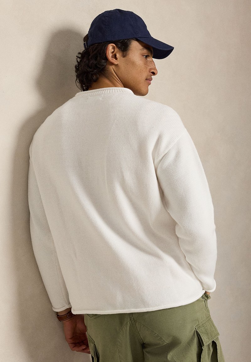 Homme portant un pull en maille blanc, un pantalon cargo vert olive et une casquette bleu marine, légèrement tourné de côté sur un fond uni.