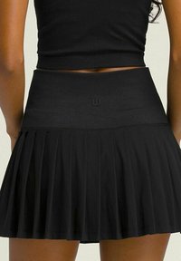 Falda plisada negra con una cinturilla alta. Hecha de tela suave, presenta un logo sutil en la cinturilla y tiene una textura suave.