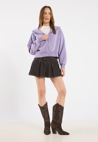 Felpa viola corta con zip frontale, abbinata a una gonna plissettata in denim nera e stivali da cowboy ricamati marroni.