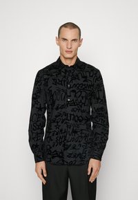 Camicetta nera con bottoni caratterizzata da un motivo testurizzato di testo e simboli stilizzati, con maniche lunghe e un classico disegno del colletto.