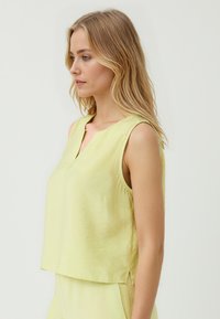 Una donna con lunghi capelli ondulati indossa un top senza maniche di un delicato giallo chiaro con un collo raffinato, guardando pensierosa di lato su uno sfondo pallido.