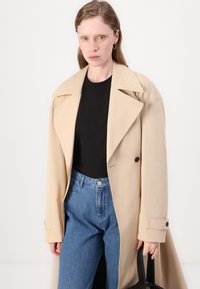 Beige trenchcoat med brede slag, dobbeltradet design og knappedetaljer, båret over en svart skjorte og blå jeans. Teksturert stoff.