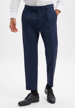 Mann trägt marineblaue Nadelstreifen-Hosen, schwarze Lederschuhe und ein weißes langärmliges Hemd, eine Hand in der Tasche, stehend.