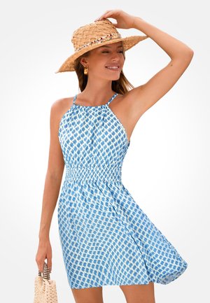 Vrouw draagt een blauw-wit patroonloze mouwloze jurk en een strohoed met brede rand, houdt een gevlochten tas vast met een glimlach, poserend tegen een effen achtergrond.