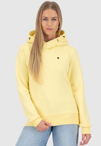 Jonge vrouw met lang blond haar, gekleed in een gele hoodie en lichtblauwe spijkerbroek, tegen een effen lichtgrijze achtergrond.