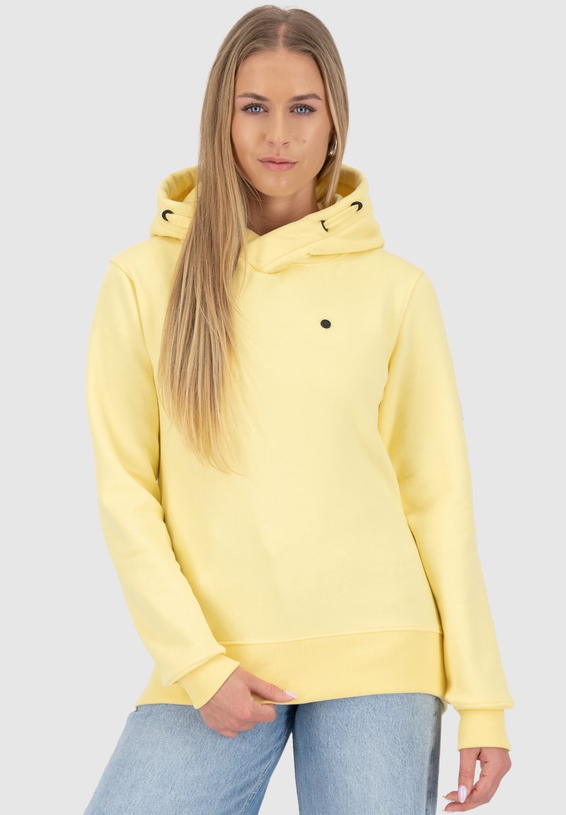 Jonge vrouw met lang blond haar, gekleed in een gele hoodie en lichtblauwe spijkerbroek, tegen een effen lichtgrijze achtergrond.
