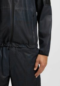Dark blue athletic jacket featuring a textured geometric pattern, zip closure, elastic waist, and contrasting black cuffs.