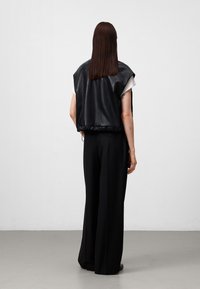 Gilet di pelle nera con una vestibilità ampia, maniche corte e un orlo leggermente arricciato, abbinato a pantaloni neri larghi e una camicia bianca sottostante.