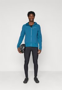 Jachetă ușoară albastră cu glugă, fermoar frontal și logo; asortată cu colanți negri pentru ciclism și pantofi. Ținând un cască de bicicletă neagră.