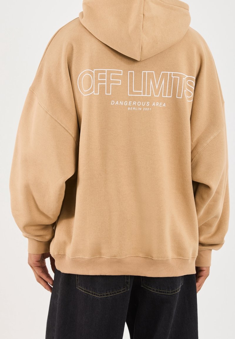 Personne portant un sweat à capuche oversize couleur sable avec l'inscription « OFF LIMITS Dangerous Area Berlin 2001 » imprimée en blanc dans le dos, associé à un jean foncé.