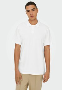 Hvid polo skjorte med ribbet tekstur, korte ærmer og en to-knaps brystlomme. En klassisk silhuet parret med lyse beige bukser.