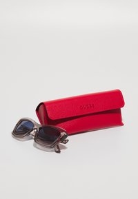 Sunglasses in transparent gray frame with dark lenses beside a red textured leather case labeled "GUESS."