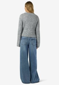 Kvinna med blont hår iklädd en grå fluffig tröja och vida blå jeans, stående med ryggen mot en enfärgad bakgrund.