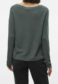 Dunkelgrüner Strickpullover mit weitem Ausschnitt, geripptem Textur an den Schultern und lockerer Passform. Kombiniert mit schwarzen Hosen.