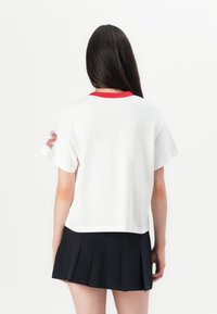 White cotton t-shirt with a red neckline and short sleeves, featuring a large "22" printed in red on the left sleeve. Paired with a black pleated skirt.