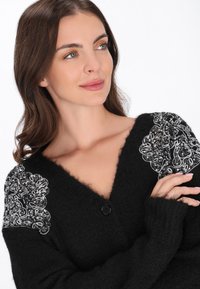 Fekete kardigán V-kivágással, texturált anyaggal és bonyolult ezüst hímzéssel a vállakon. Egy gombos zárás elöl.