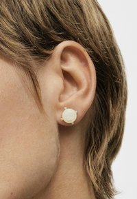 Pendientes de cerámica blanca con acentos plateados, que presentan una superficie lisa y una forma redonda, asegurados con pequeños detalles decorativos.