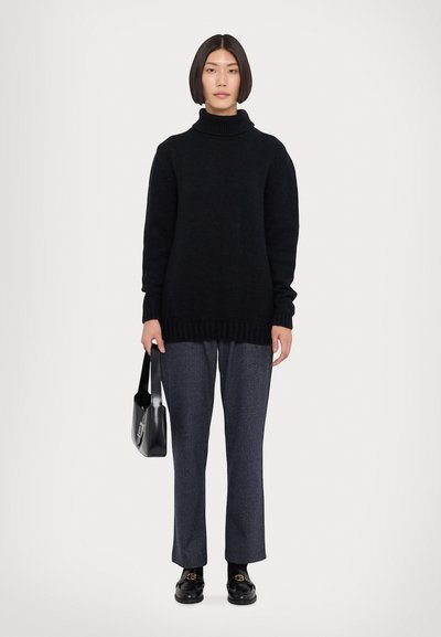 Svart strikket turtleneck genser, løs passform, ribbestrikkede mansjetter og kant. Kombinert med grå skreddersydde bukser og svarte loafers, holder en svart håndveske.
