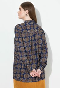 Blusa a fantasia navy con disegni intricati in oro e marrone, maniche lunghe e vestibilità morbida. La vista posteriore mostra un collo con bottoni.