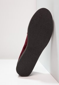 Chaussure en daim bordeaux avec une semelle en caoutchouc noir texturé, dotée d'un bout arrondi et de bords lisses, inclinée contre un fond blanc.