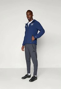 Granatowa kurtka przeciwwiatrowa z zamkiem z przodu i kapturem, z naszywką. W połączeniu z szarymi spodniami sportowymi i czarnymi butami sportowymi, prezentującymi smukły krój.