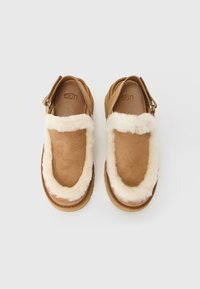Een paar tan suède UGG-slippers met een witte fluffy voering en een verstelbare achterkant op een witte achtergrond.
