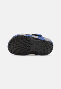 Black rubber shoe sole with a textured pattern, featuring blue accents and a grey woven upper. Fastening strap located on the side.