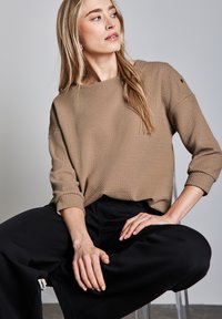 Beiger strukturierter Pullover mit lockerer Passform, rundem Halsausschnitt und umgeschlagenen Ärmelbündchen, kombiniert mit schwarzen weit geschnittenen Hosen. Klare Hintergrund.