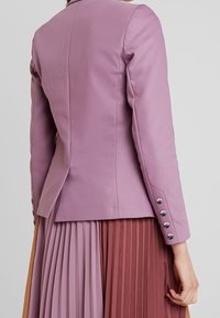 Lawendowa marynarka o dopasowanym kroju, z czterema srebrnymi guzikami na każdym rękawie i eleganckim tyłem. Plisowana spódnica w uzupełniających się kolorach.