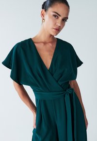 Green wrap dress in lightweight fabric, featuring a deep V-neck, short sleeves, and a tied waist. Smooth texture with minimal design accents.
