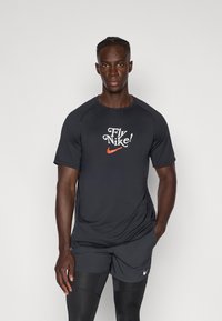 Fekete sportpóló kerek nyakkivágással, fehér és narancssárga felirattal: "Fly Nike!". Rövid raglán ujjakkal rendelkezik és sima tapintású.