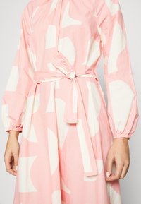 Růžové šaty s bílým abstraktním vzorem, dlouhé rukávy s elastickými manžetami a detaily s vázáním v pase. Vyrobeno z lehkého materiálu.