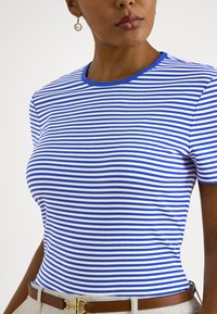 Γυναίκα που φορά μπλε και άσπρη ριγέ μπλούζα, μπεζ παντελόνι με καφέ ζώνη και χρυσό κυκλικό σκουλαρίκι στο αριστερό αυτί, εμφανίζεται από το στήθος και πάνω.