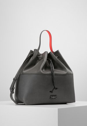 Sac seau en cuir noir avec moitié inférieure texturée, moitié supérieure lisse, fermeture à cordon et anse unique avec-dessous rouge sur un pied blanc.