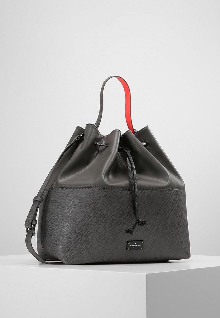 Sac seau en cuir noir avec moitié inférieure texturée, moitié supérieure lisse, fermeture à cordon et anse unique avec-dessous rouge sur un pied blanc.