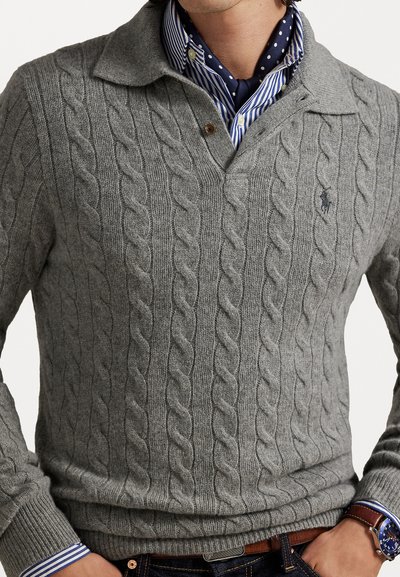 Pull en tricot gris à col, affichant un logo discret, porté sur une chemise rayée bleue et blanche et un jean en denim foncé.